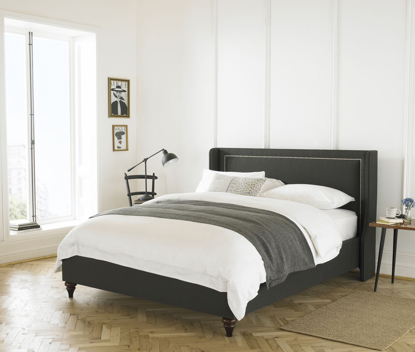 Charcoal upholstered bed with nailhead trim styled beneath framed Vogue prints on a herringbone floor