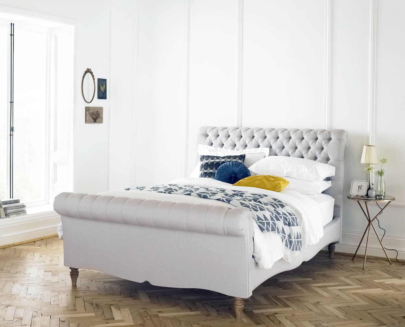 Pale grey button-tufted chesterfield sleigh bed styled in a bedroom with navy and mustard cushions